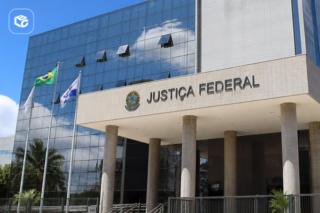 Edifício moderno da Justiça Federal com fachada de vidro espelhado refletindo nuvens no céu azul. Na entrada, colunas altas sustentam a cobertura, onde estão visíveis o brasão da República e a inscrição “JUSTIÇA FEDERAL”. Três mastros com bandeiras, incluindo a bandeira do Brasil, estão posicionados em frente ao prédio.