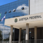 Edifício moderno da Justiça Federal com fachada de vidro espelhado refletindo nuvens no céu azul. Na entrada, colunas altas sustentam a cobertura, onde estão visíveis o brasão da República e a inscrição “JUSTIÇA FEDERAL”. Três mastros com bandeiras, incluindo a bandeira do Brasil, estão posicionados em frente ao prédio.